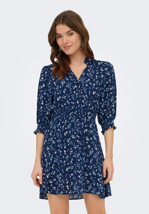 Jeune femme aux cheveux bruns ondulés portant une robe bleu marine à imprimé floral avec des manches trois-quarts et une taille cintrée, debout devant un fond uni.