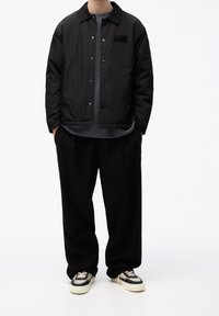 Czarna nylonowa kurtka na zatrzaski z kołnierzem i logo na piersi, zestawiona z luźnymi czarnymi spodniami i czarno-białymi sneakersami.