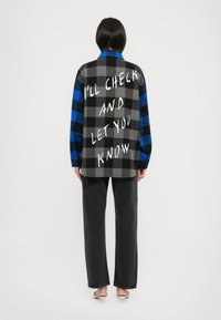 Camicia oversize a quadri blu e nera con retro grigio a scacchi caratterizzato da testo bianco. Abbinata a pantaloni scuri e calzature chiare.
