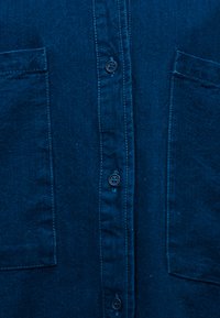 Σκούρο μπλε ύφασμα με ορατή ραφή, κάθετη σειρά κουμπιών με σκούρα στρογγυλά κουμπιά και δύο ορθογώνιες τσέπες επάνω στο ύφασμα.