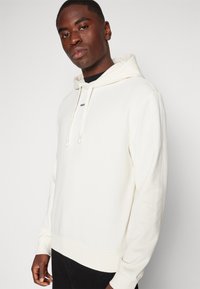 Svijetlosmeđa hoodie s kapuljačom, mekane teksture, s rebrastim manžetama i rubovima; ima suptilan logo brenda na prednjoj strani.