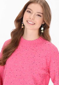 Rózsaszín pulóver kerek nyakkal, színes, apró fémes szórásokkal. Hosszú haj és csillagformájú fülbevalók díszítik.