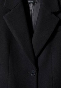 Manteau en laine foncée avec de larges revers et un seul bouton noir fixé près du centre devant.