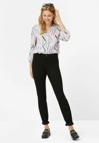 Bluse mit mehrfarbigem geometrischem Muster und langen Ärmeln, kombiniert mit engen schwarzen Jeans und schwarzen Loafers mit goldenen Hardware-Akzenten.