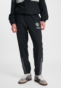 Svarta träningbyxor med elastisk midja, grå sidopaneler och en logotyp på låret, matchade med grå sneakers med marinblå detaljer.