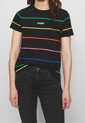 Frau trägt schwarzes Wrangler-T-Shirt mit horizontalen Regenbogenstreifen und schwarze Jeans, steht vor hellem Hintergrund.