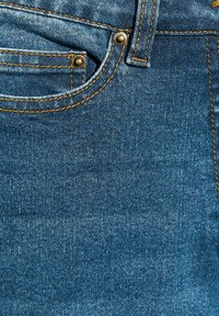 Tejido de denim azul oscuro que muestra un primer plano de un bolsillo, con costuras doradas y remaches de metal, presentando una superficie texturizada.