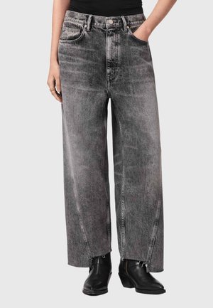 Person iført højtsiddende, vidbenede, falmede sorte jeans med rå kant og sorte ankelstøvler, hænderne i forlommerne, mod neutral baggrund.