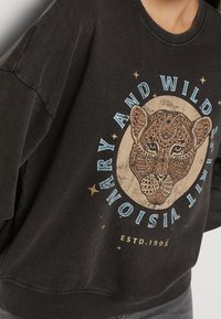 Černá mikina s velkým, detailně zpracovaným grafickým vyobrazením leopardího obličeje s složitými vzory a modrým textem "VIZE A DIVOKÝ DUCH."