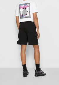 T-shirt blanche à manches courtes avec impression graphique au dos, accompagnée de shorts noirs et de chaussures noires avec des accents verts et un design texturé.