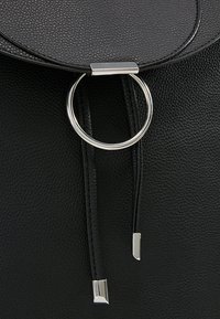 Sac en cuir noir texturé avec un anneau circulaire argenté et deux lanières en cuir suspendues avec des embouts en métal argenté sous le rabat.