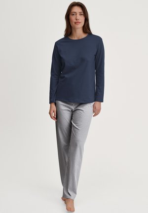 Vrouw staat blootsvoets en draagt een donkerblauw shirt met lange mouwen en een witte broek met dunne verticale zwarte strepen tegen een effen achtergrond.
