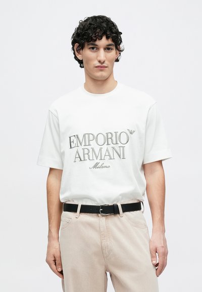 Biele bavlnené tričko s krátkymi rukávmi, na prednej strane šedý potlač s nápisom "EMPORIO ARMANI Milano." Model má na sebe béžové nohavice a čierny opasok.