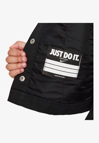 Interior de chaqueta negra que muestra una etiqueta con el texto "JUST DO IT." en blanco negrita, junto con cierres de presión y textura acolchada.