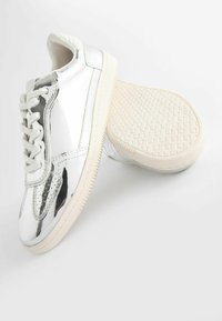 Sneaker in vernice argento con suole bianche, caratterizzati da un design con lacci, dettagli perforati e una suola in gomma testurizzata.