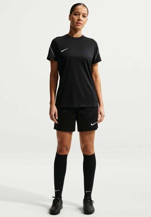 Mujer de pie vistiendo ropa deportiva negra de Nike que incluye camiseta, pantalones cortos, calcetines largos y botas de fútbol sobre un fondo liso y claro.