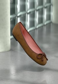 Ballet flat en suède marron avec un intérieur rose, bout arrondi, détail de nœud décoratif et une semelle noire basse. Design minimaliste avec une texture lisse.