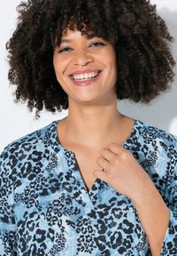 Blaue Bluse mit Leopardenmuster, V-Ausschnitt und lässiger Passform. Sie hat schwarze Flecken auf einem hellblauen Hintergrund, eine weiche Textur und dreiviertellange Ärmel.