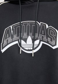 Czarna bluza z kapturem z białym logo „ADIDAS” oraz grafiką koniczyny. Materiał jest miękki, o prążkowanej fakturze, z regulowanymi sznurkami zakończonymi metalowymi zakończeniami.
