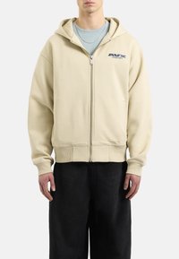 Felpa beige con zip, cappuccio e tasche a marsupio, con logo sul petto sinistro. Il tessuto strutturato e i polsini a coste completano il design.