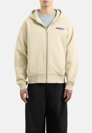 Sudadera cremosa con cremallera, capucha y bolsillos tipo canguro, con un logo en el lado izquierdo del pecho. El diseño se completa con un tejido texturizado y puños acanalados.