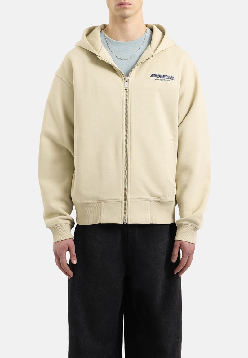 Felpa beige con zip, cappuccio e tasche a marsupio, con logo sul petto sinistro. Il tessuto strutturato e i polsini a coste completano il design.