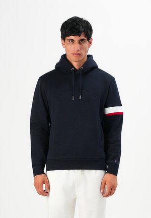 Homme portant un sweat à capuche bleu marine avec une bande blanche et rouge sur le bras droit et un pantalon blanc, se tenant devant un fond blanc.