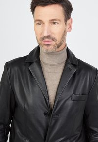 Czarna skórzana marynarka z pojedynczym zapięciem na guzik, wyciętymi klapami i kieszenią na piersi, noszona na jasnobeżowym swetrze z golfem.