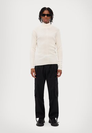 Crème ribbeltrui met hoge Hals, gecombineerd met zwarte cargobroek. Zwarte zonnebril en chunky zwarte schoenen maken de look compleet.
