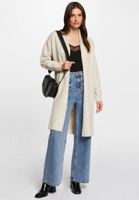 Ljust beige lång kofta i mjuk ullblandning, bärs över en svart topp med spetsdetaljer. Matchas med högmidjade blå vida jeans och svarta klackar.