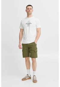 Camiseta de algodón blanca con texto verde, combinada con pantalones cortos de cargador verde oliva con bolsillos laterales. Zapatillas marrones con detalles en blanco.
