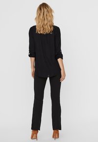 Blusa nera con maniche lunghe, arrotolate fino ai gomiti, in tessuto morbido, abbinata a pantaloni neri svasati e scarpe con tacco marroni.