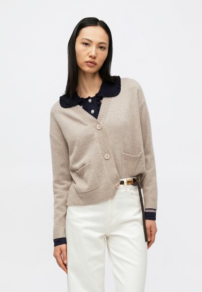 Frau trägt eine beige geknöpfte Strickjacke über einem marineblauen Hemd mit gerafftem Kragen, weiße Hose und braunen Gürtel, steht vor einem schlichten hellen Hintergrund.