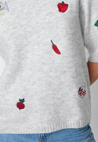 Maglione lavorato a maglia grigio chiaro con piccolo peperoncino rosso, peperone, barbabietola e un cestino da picnic ricamati, indossato con jeans blu.