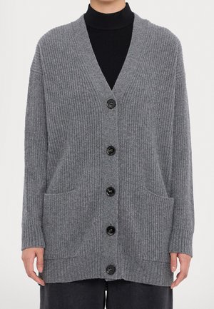 Person wearing a grey ribbed knit cardigan with black buttons and front pockets over a black high-neck top.