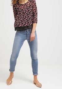 Blouse noire à fleurs avec manches trois-quarts, associée à un jean skinny bleu clair et des ballerines métalliques. Style décontracté et cintré.