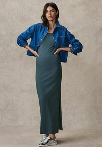 Maxi dress verde a coste con uno spacco laterale, abbinata a una giacca di denim blu. Il modello indossa sneakers argentate. Sfondo neutro e testurizzato.
