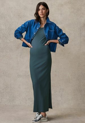 Grön räfflad maxiklänning med en sidoklyfta, kombinerad med en blå denimjacka. Modellen bär silversneakers. Neutral texturerad bakgrund.