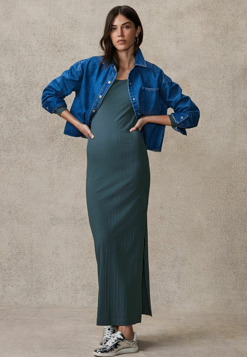 Maxi dress verde a coste con uno spacco laterale, abbinata a una giacca di denim blu. Il modello indossa sneakers argentate. Sfondo neutro e testurizzato.