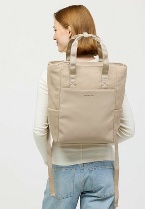 Frau mit roten Haaren, die einen cremefarbenen Pullover und blaue Jeans trägt und einen beigen Rucksack mit Vorderfach und oberen Griffen auf dem Rücken hat.