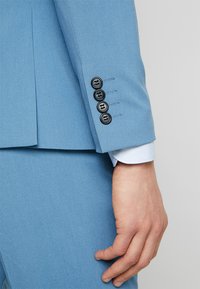 Manga de chaqueta de traje azul claro que presenta cuatro botones negros y detalles de costura ajustada. Tejido suave con un diseño estructurado.
