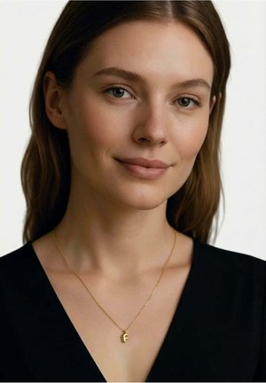 Junge Frau mit hellem Teint, glatten braunen Haaren, trägt ein schwarzes Oberteil und eine goldene Halskette mit Anhänger in Form des Buchstabens "F", lächelt sanft.