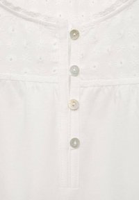 Blusa bianca con motivi floreali ricamati, scollo rotondo e cinque bottoni iridescenti lungo il davanti. Tessuto di cotone morbido.