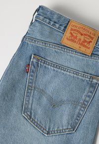 Φωτεινό μπλε τζιν από denim διπλωμένο για να φαίνεται η πίσω τσέπη με κίτρινη ραφή, κόκκινη ετικέτα Levi's και ταμπέλα από δέρμα Levi Strauss σε καφέ χρώμα.