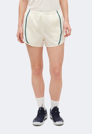 Άτομο που φοράει λευκά αθλητικά σορτς με πράσινες πλαϊνές ρίγες, λευκές κάλτσες και μαύρα παπούτσια τρεξίματος, στέκεται μπροστά σε φωτεινό φόντο.