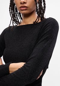 Maglietta nera a maniche lunghe in tessuto testurizzato e lucente. Presenta un ampio scollo e una silhouette aderente, con maniche lunghe e senza hardware visibile.