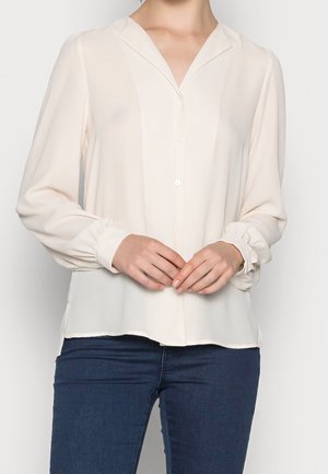 Blusa crema con scollo a V, maniche lunghe e polsini arricciati. Bottoni sul davanti; tessuto liscio e leggero con spacchi laterali all'orlo.