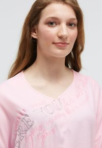 Young woman with long light brown hair wearing a pink shirt with faded self-reflection and release-themed text.