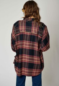 Chemise à carreaux surdimensionnée en noir, rouge et beige avec des manches courtes. Présente des clous en métal sur les épaules et le dos pour un détail supplémentaire.