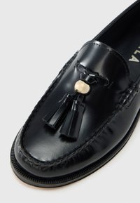 Μαύρα δερμάτινα μοκασίνια με στρογγυλεμένη μύτη, διακοσμημένα με φούντες και λεπτομέρεια από χρυσό κουμπί. Λείο υλικό με ραμμένες άκρες.